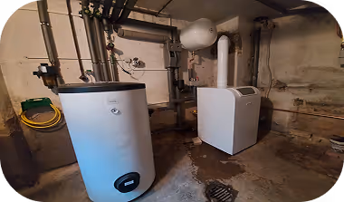 Keller mit Heizungsanlage, darunter ein runder Boiler und eine weiße Heizungs- oder Lüftungseinheit, mit sichtbaren Rohrleitungen und einem Abfluss im Boden.