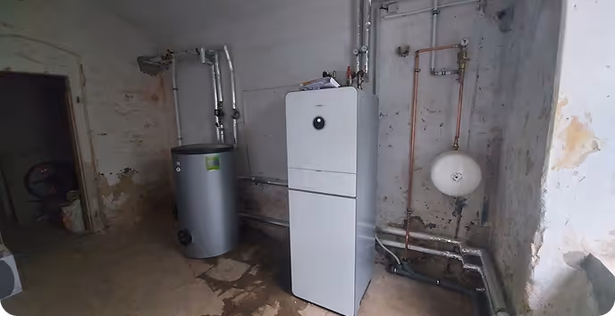 Heizungsanlage mit Boiler in einem Kellerraum mit sichtbaren Rohrleitungen und abblätternden Wänden.