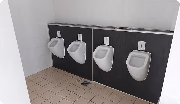 Vier weiße Urinale an einer dunkelgrauen Wand in einer öffentlichen Toilette mit beigen Bodenfliesen.