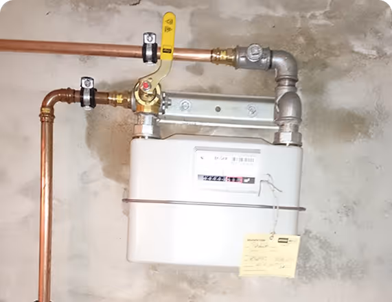 Gaszähler mit Kupferrohren und gelbem Absperrventil an einer Wand montiert.