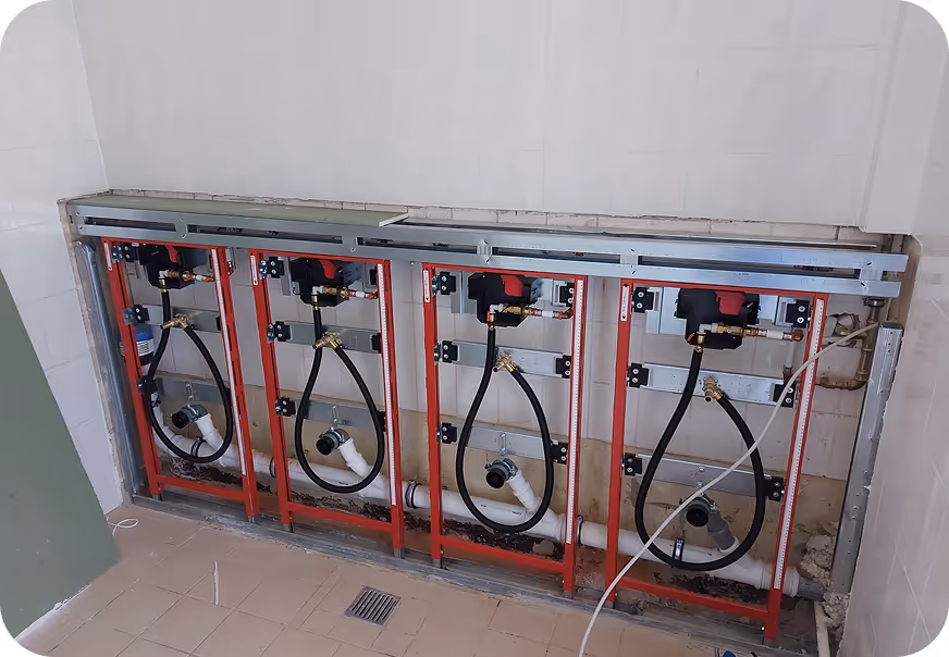 Rohinstallation für vier Wand-WC-Elemente mit rotem Metallrahmen und schwarzen Wasseranschlüssen in einem unfertigen Badezimmer.