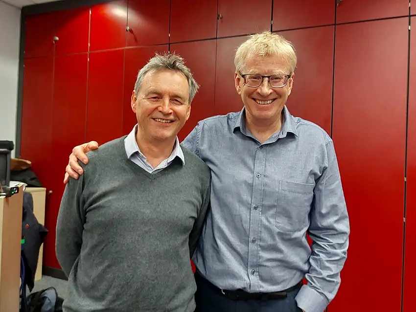 Two middle-aged men smiling, one with grey hair and a grey sweater, the other with blonde hair and glasses wearing a blue shirt, standing in front of red cabinets.
