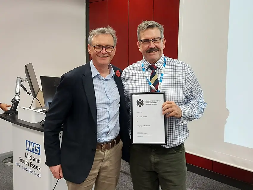 Two men smiling and posing for a photo in an office setting; one holds a framed certificate and both wear glasses.