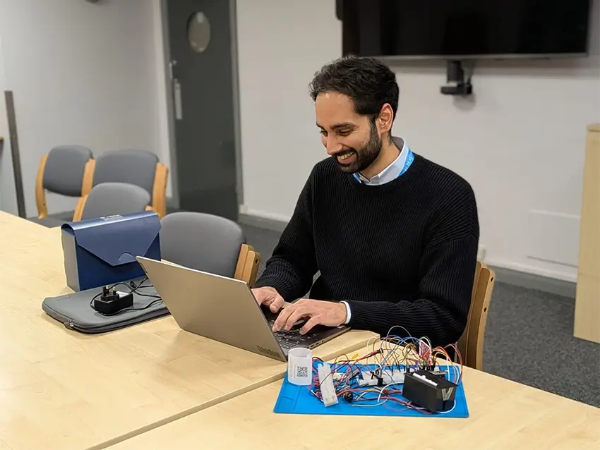 Man smiling while working on a laptop next to an electronics breadboard with wires and components on a wooden table.