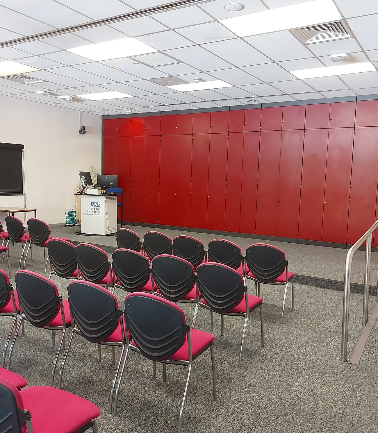 Empty seminar room with rows of pink and black chairs facing a small stage with a podium against a red paneled wall.