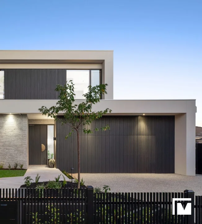 Modern two-story house in Malvern with grey vertical paneling, a large garage door, and a small tree in the front yard under a clear blue sky.