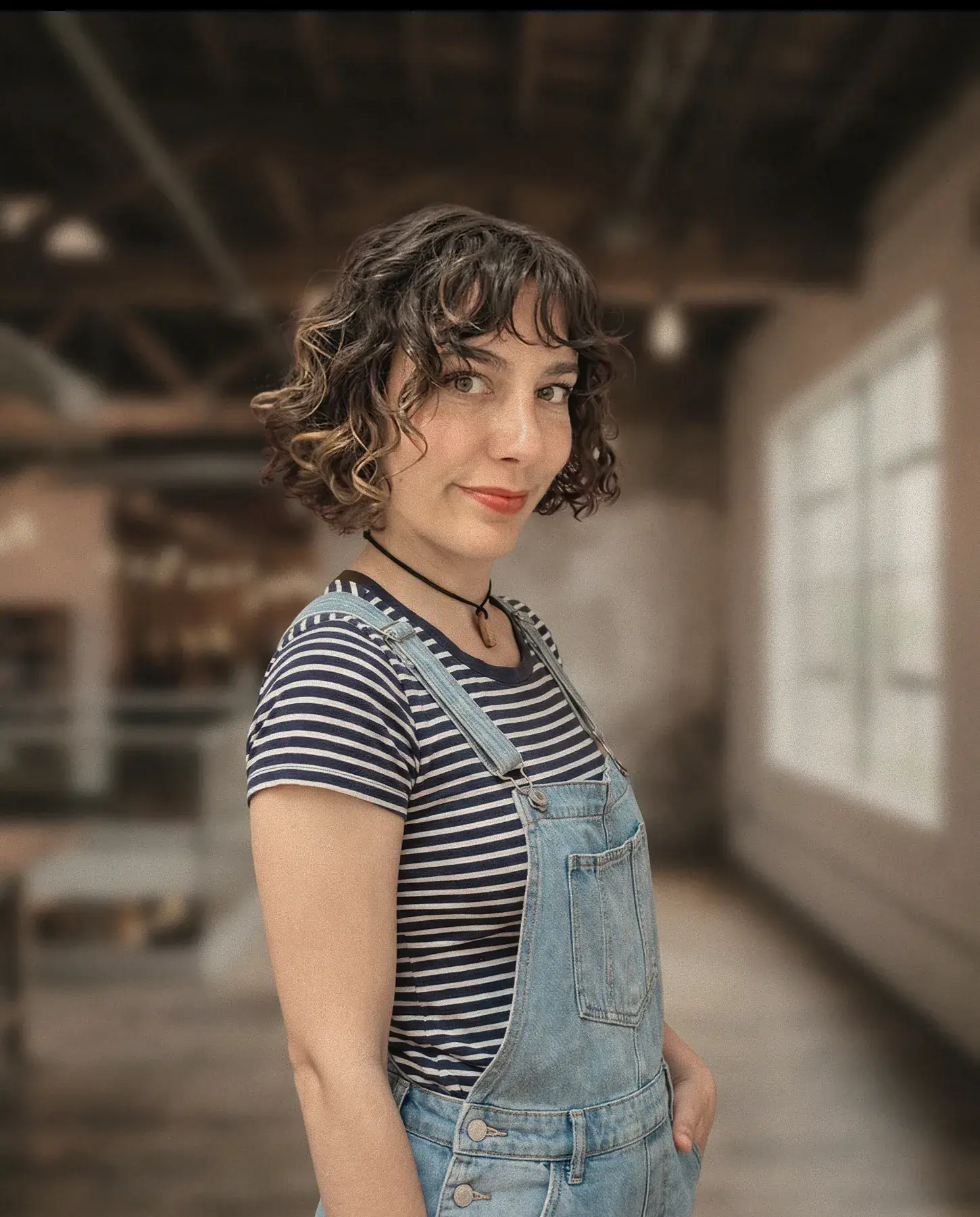 Femme aux cheveux bouclés courts avec mèches claires, portant un t-shirt rayé noir et blanc sous une salopette en jean, souriant légèrement, posant dans un espace intérieur flou avec une grande fenêtre.