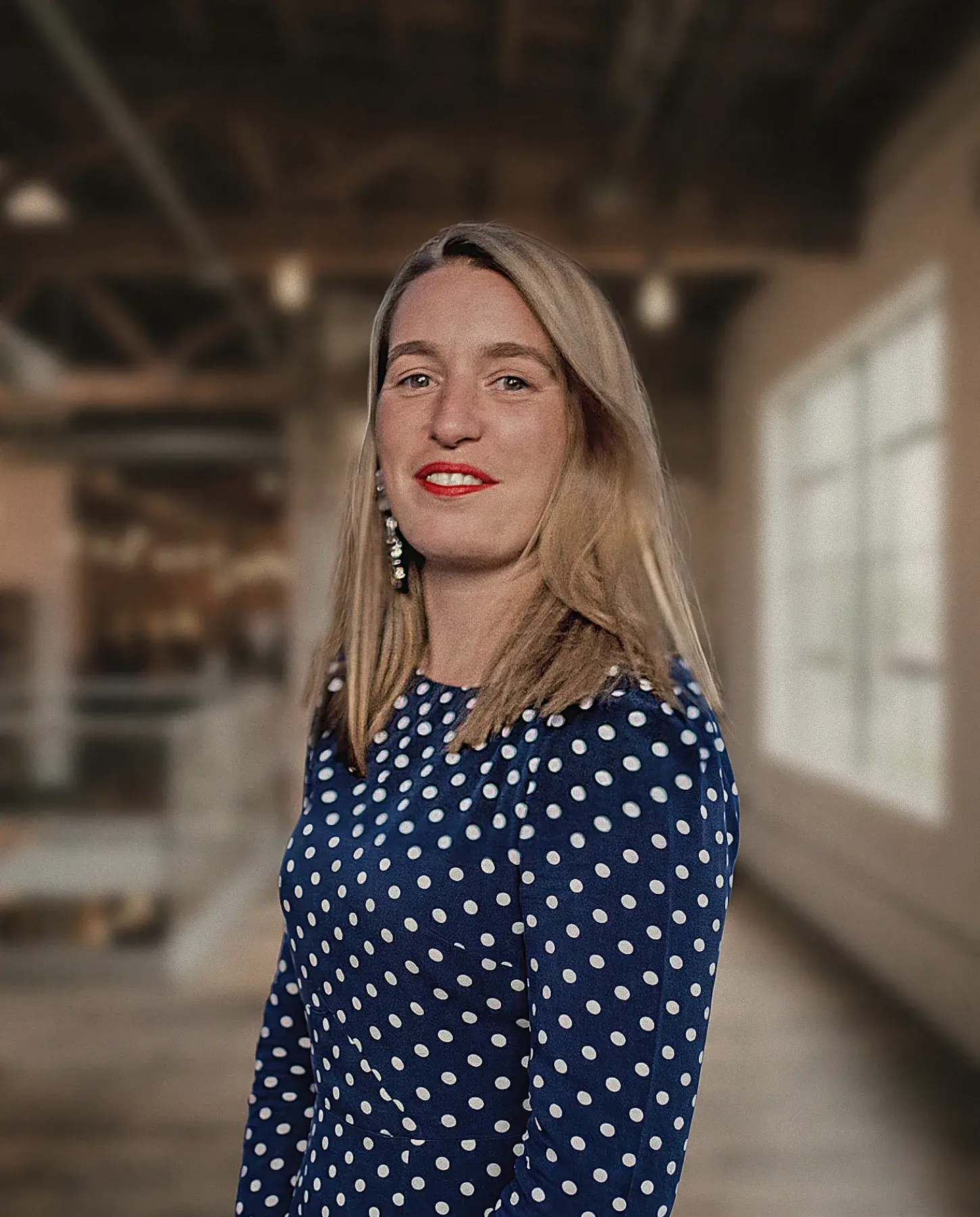 Femme aux cheveux blonds mi-longs portant une robe bleue à pois blancs avec boucles d'oreilles pendantes, souriant dans un espace intérieur flou avec grandes fenêtres.