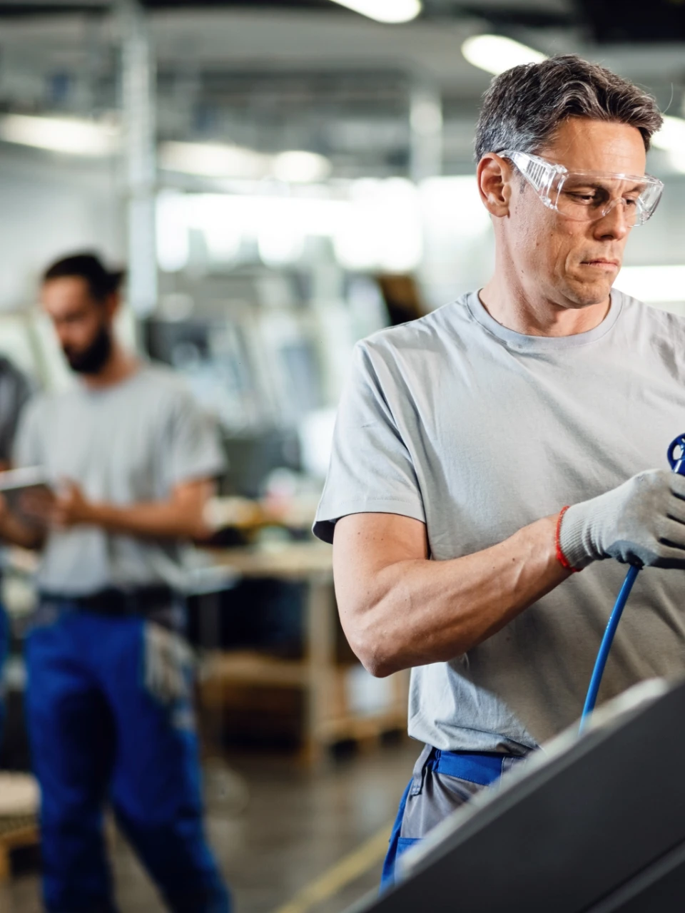 Industrieller Arbeiter mit Schutzbrille und Handschuhen bedient in einer Fabrik eine Maschine, im Hintergrund ein weiterer Arbeiter.