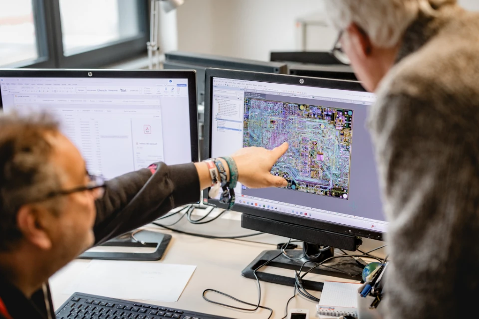 Zwei Männer betrachten und diskutieren ein Leiterplattendesign auf einem Computermonitor in einem Büro.