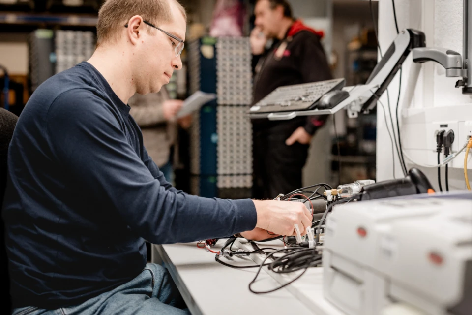 Ein Mann mit Brille arbeitet konzentriert an elektronischen Geräten und Kabeln auf einem Tisch in einem Labor oder Werkstatt.