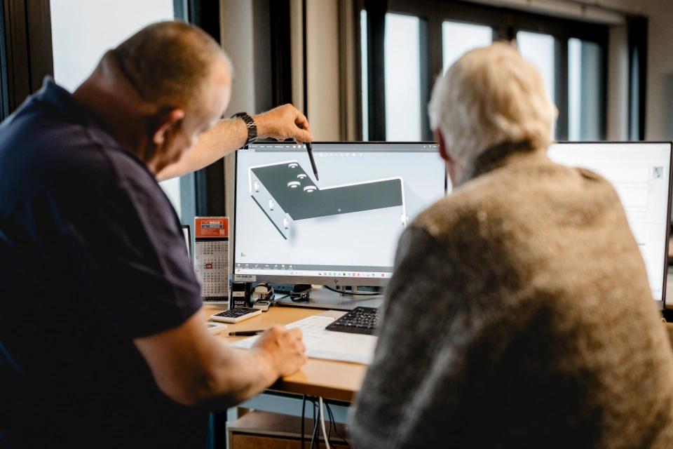Zwei Männer sitzen an einem Schreibtisch und betrachten eine 3D-Konstruktion eines Metallteils auf einem Computerbildschirm.