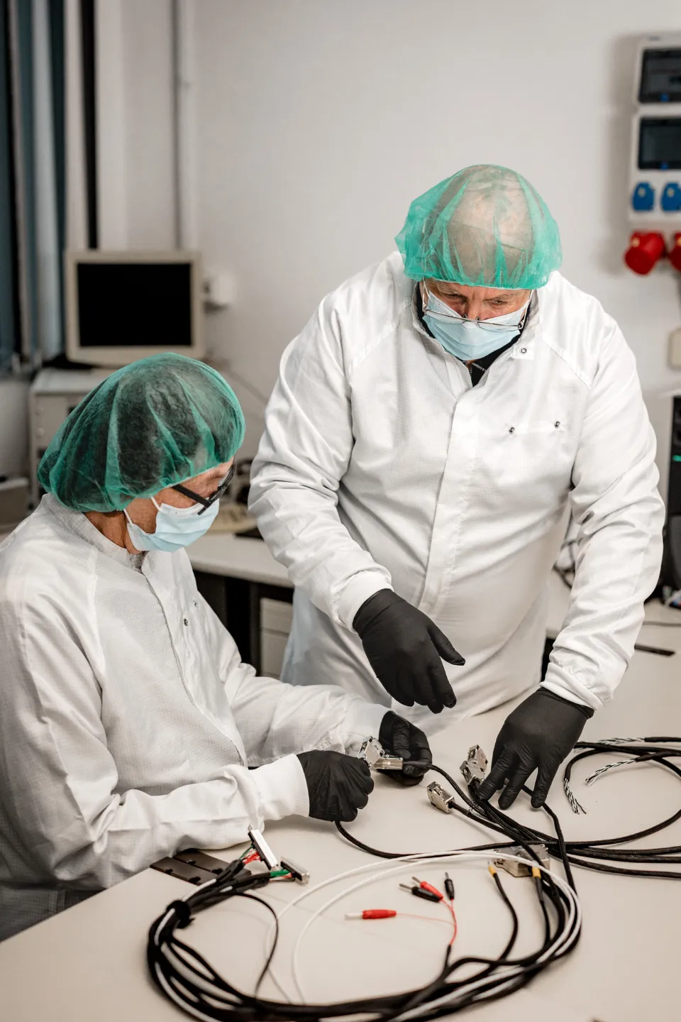 Zwei Personen in weißen Schutzanzügen, grünen Haarnetzen und Gesichtsmasken bearbeiten elektrische Kabel an einem Labortisch.