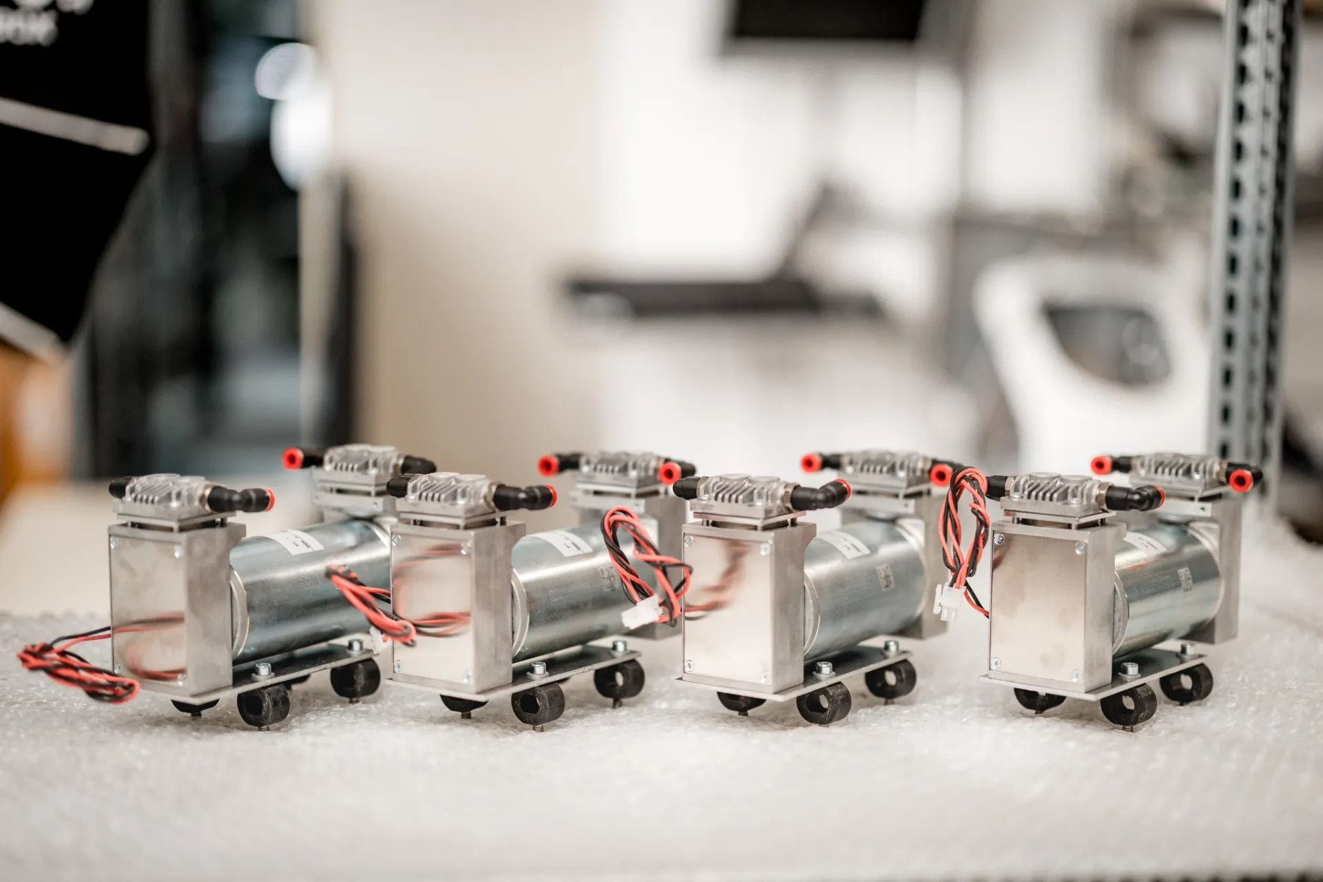 Vier kleine metallische Luftkompressoren mit angeschlossenen roten und schwarzen Kabeln auf einer gepolsterten Oberfläche.