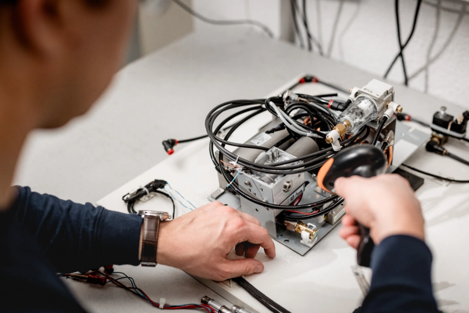 Person hält einen Barcode-Scanner über ein medizinisches Gerät mit Kabeln auf einem Tisch.