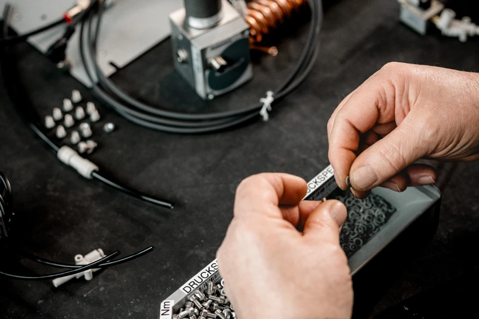 Hände sortieren kleine Metallringe auf einem Tisch mit technischen Bauteilen und Kabeln.