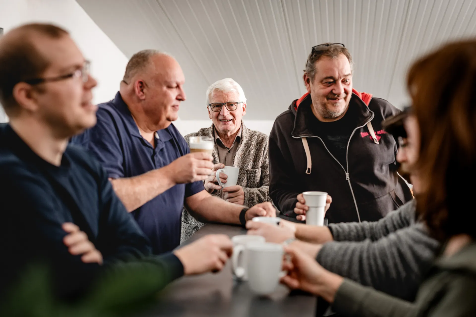 Fünf Personen unterschiedlicher Altersgruppen stehen an einem Tresen und trinken zusammen Kaffee und Bier.
