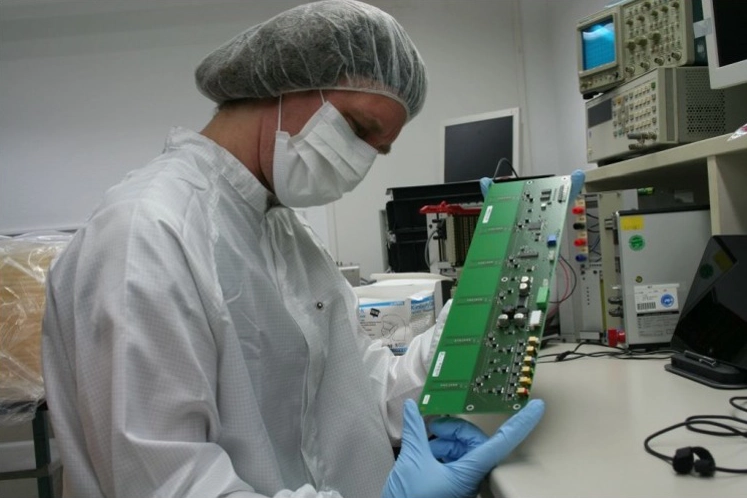 Techniker in weißem Kittel, Haarnetz, Maske und blauen Handschuhen untersucht eine grüne Leiterplatte in einem Labor mit elektronischen Messgeräten.