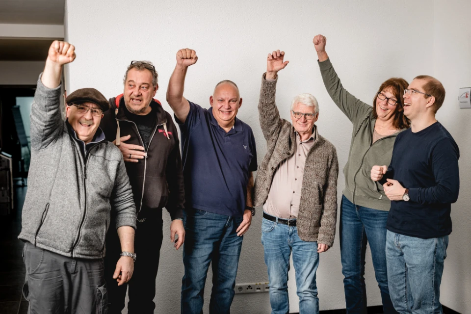 Sechs fröhliche Menschen unterschiedlichen Alters stehen vor einer weißen Wand und heben jeweils eine Faust in die Luft.