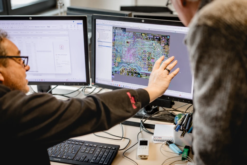 Zwei Personen diskutieren ein komplexes Schaltplan-Layout auf einem Computerbildschirm in einem Büro.
