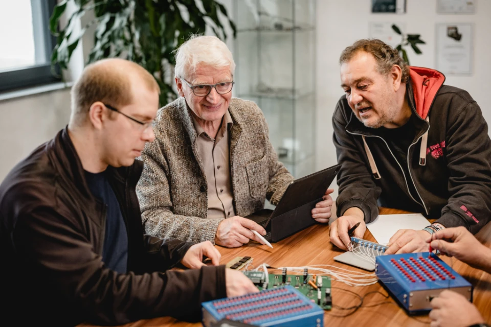 Drei Männer unterschiedlicher Generationen sitzen an einem Tisch und arbeiten gemeinsam an elektronischen Schaltungen mit blauem Steckboard und Tablet.