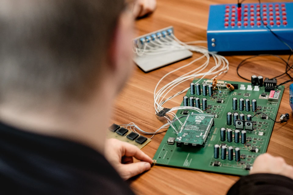 Person arbeitet an einem grünen elektronischen Schaltkreis mit vielen Bauteilen und angeschlossenen weißen Kabeln auf einem Holztisch.