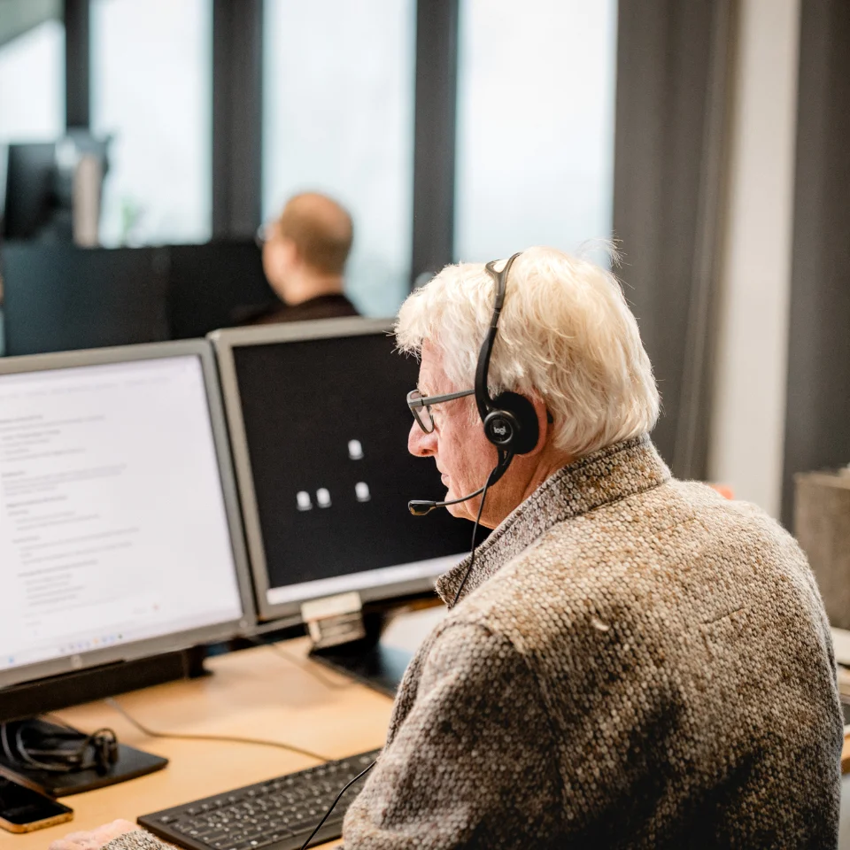Älterer Mann mit Headset arbeitet an einem Schreibtisch mit zwei Monitoren in einem Büro.