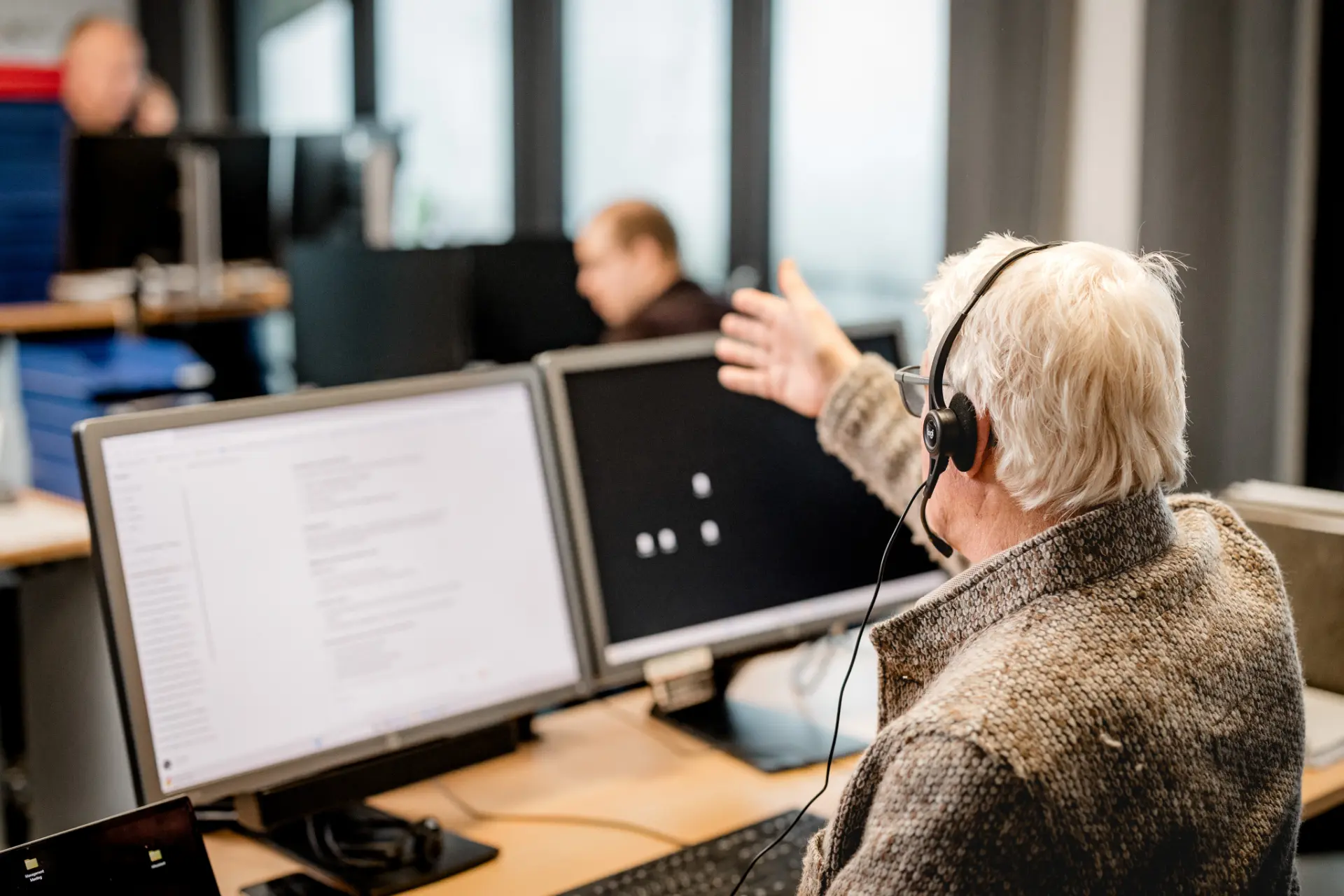 Älterer Mann mit Headset gestikuliert in einem Büro mit mehreren Computermonitoren.