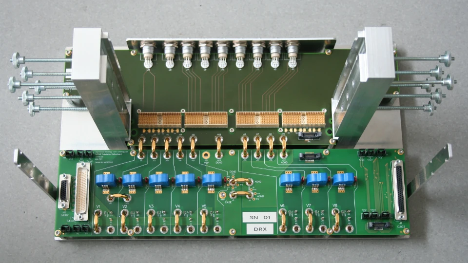 Grüne Leiterplatte mit mehreren blauen elektronischen Bauteilen, goldenen Verbindungsleitungen und zwei großen Metallklemmen an den Seiten.