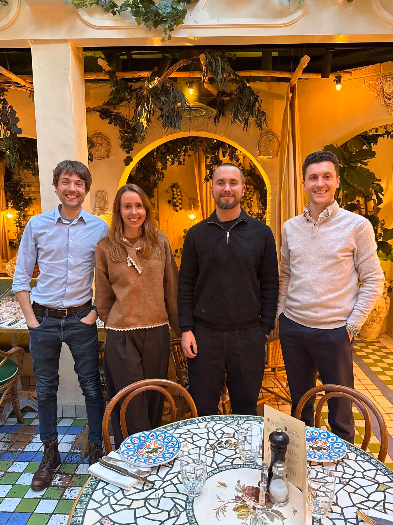 Four people smiling and standing behind a mosaic-style round table set with plates and glasses in a warmly lit restaurant with greenery and decorative arches.
