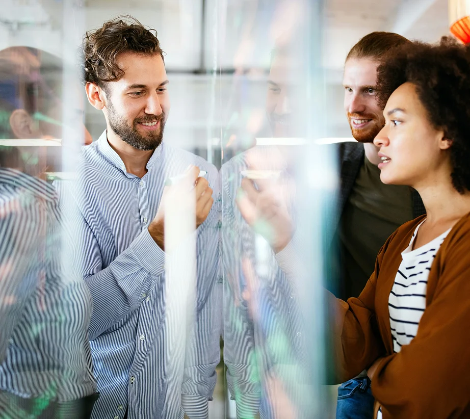 Three colleagues collaboratively discussing ideas and writing on a transparent board in an office.