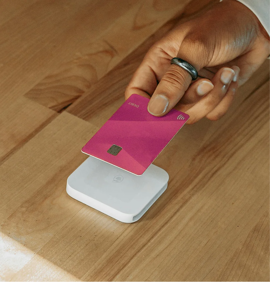 Hand holding a pink debit card above a white contactless card reader on a wooden surface.