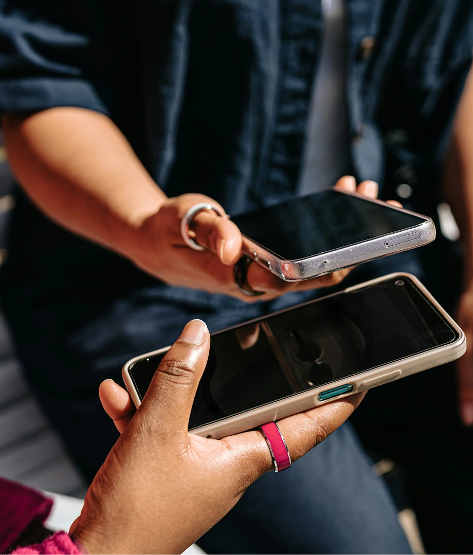 Two people holding smartphones with black screens, one hand wearing a pink ring and the other wearing silver rings.