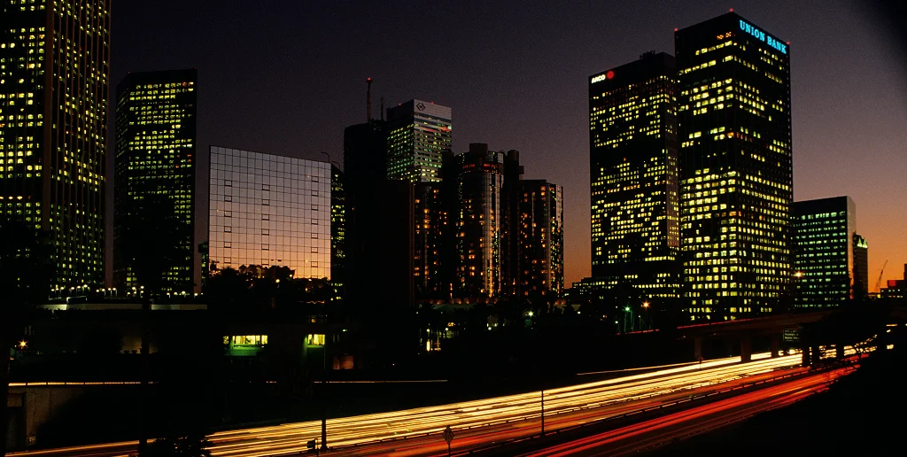 City skyline at dusk with illuminated office buildings and light trails from cars on a highway.