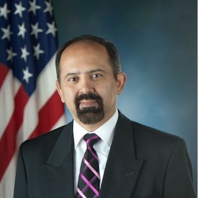 Man in suit and patterned tie posing in front of a U.S. flag and blue background.