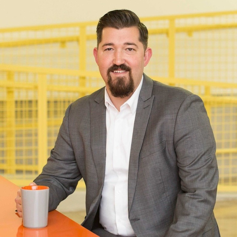 Man with goatee wearing gray suit and white shirt, sitting at orange table holding a gray mug.