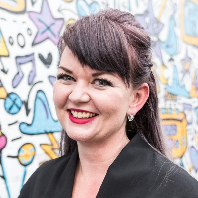 Smiling woman with dark hair and red lipstick wearing black jacket in front of colorful graffiti wall.