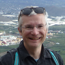 Smiling man with glasses and gray hair outdoors with a mountainous landscape background.