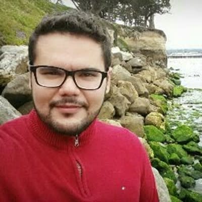 Man with black glasses and red sweater standing near rocky coastline covered in green algae.
