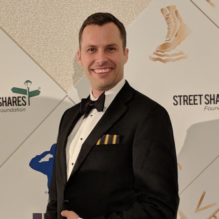 Smiling man in black tuxedo with bow tie and striped pocket square at an event backdrop.