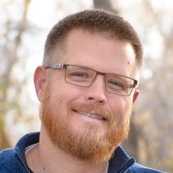 Man with short hair, glasses, and reddish beard smiling outdoors with blurred background.
