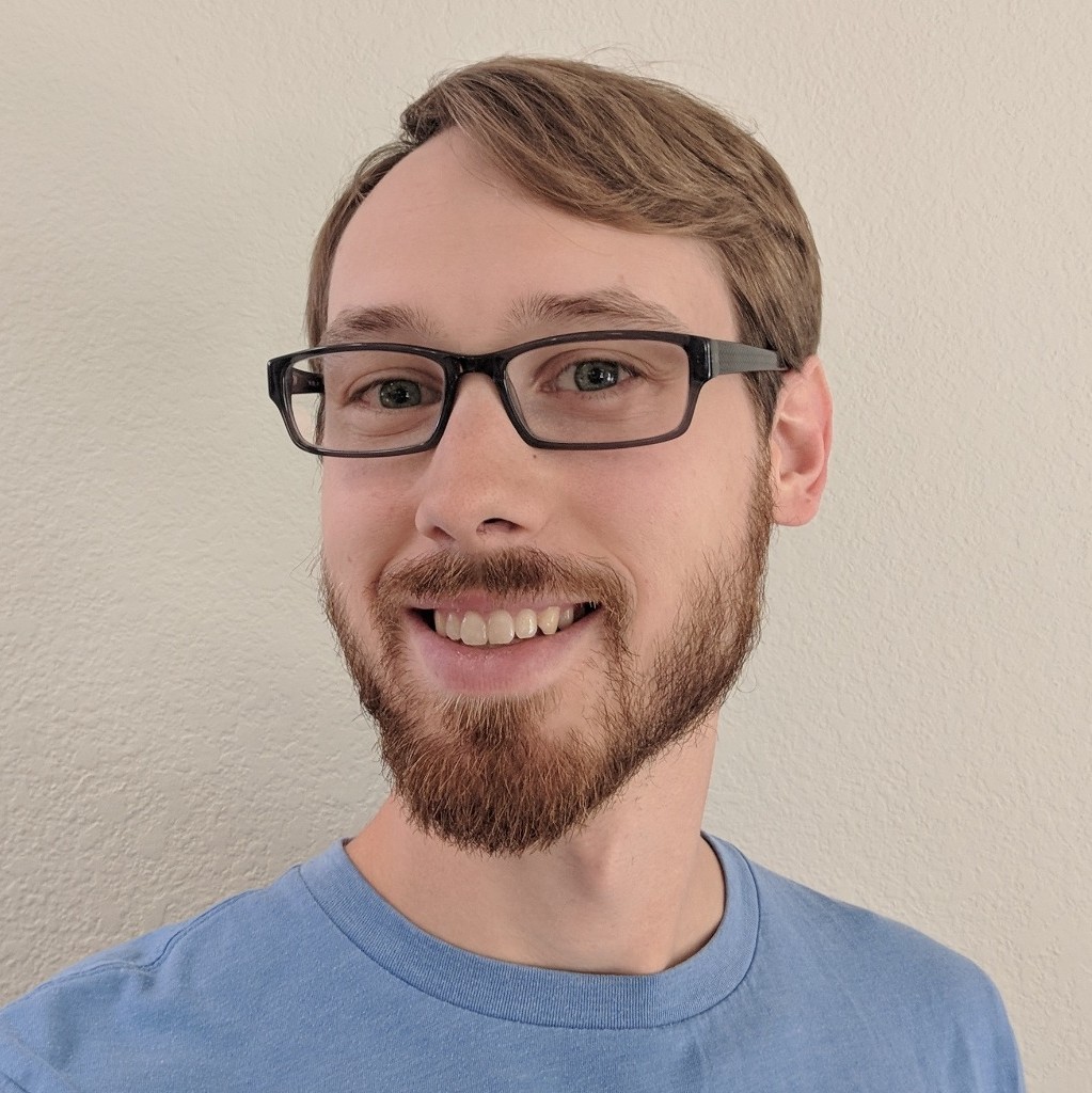 Smiling man with glasses, light brown hair, beard, and blue shirt against a beige wall.