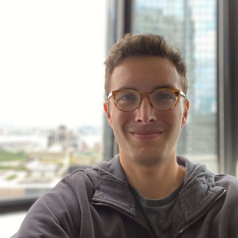 Young man with glasses and short curly hair smiling indoors by a window with city buildings outside.