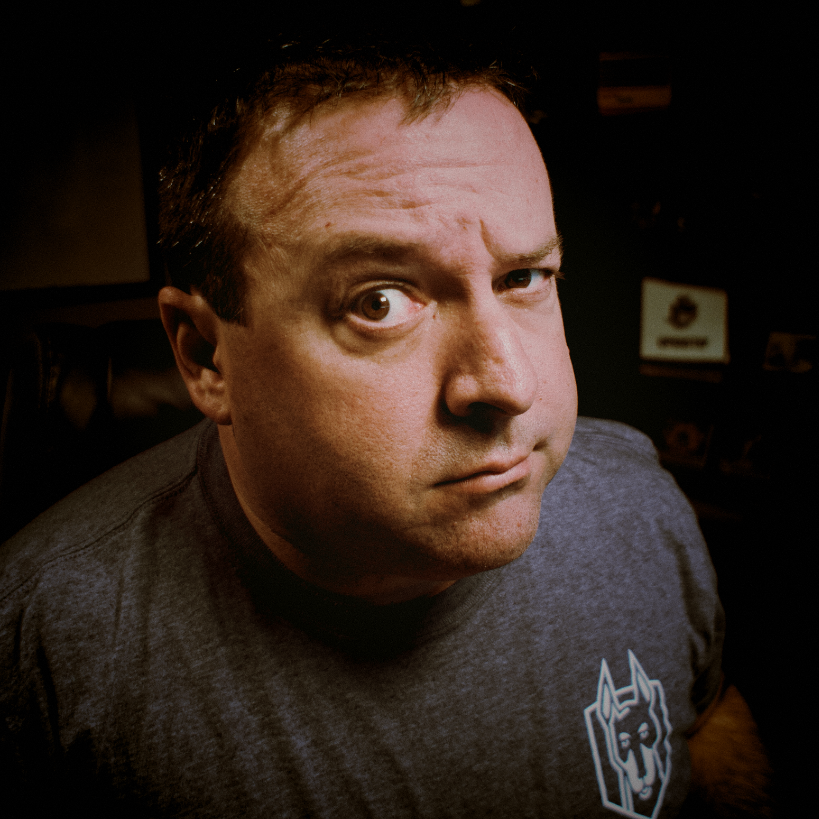 Man with short dark hair wearing a gray shirt with a dog logo, looking skeptically at the camera.