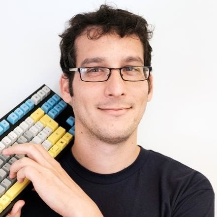 Man with glasses holding a colorful mechanical keyboard, smiling at the camera.