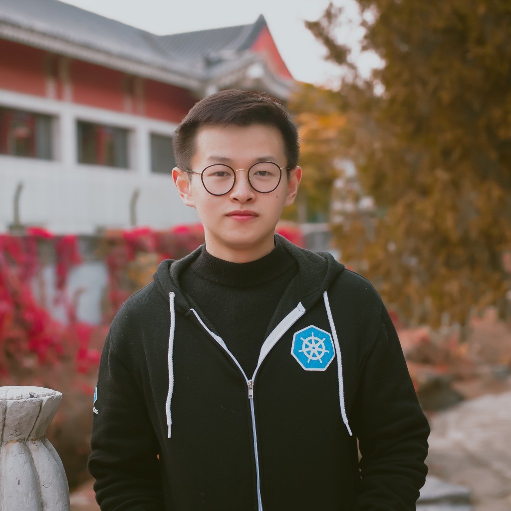 Young man with round glasses wearing black Kubernetes hoodie standing outdoors near autumn foliage.