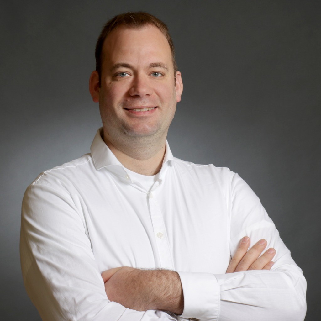 Smiling man in white button-up shirt with arms crossed against a gray background.