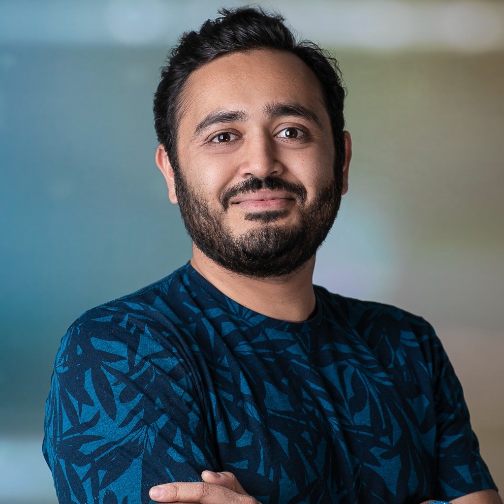 Smiling man with dark hair and beard wearing a dark blue patterned t-shirt against a blurred background.