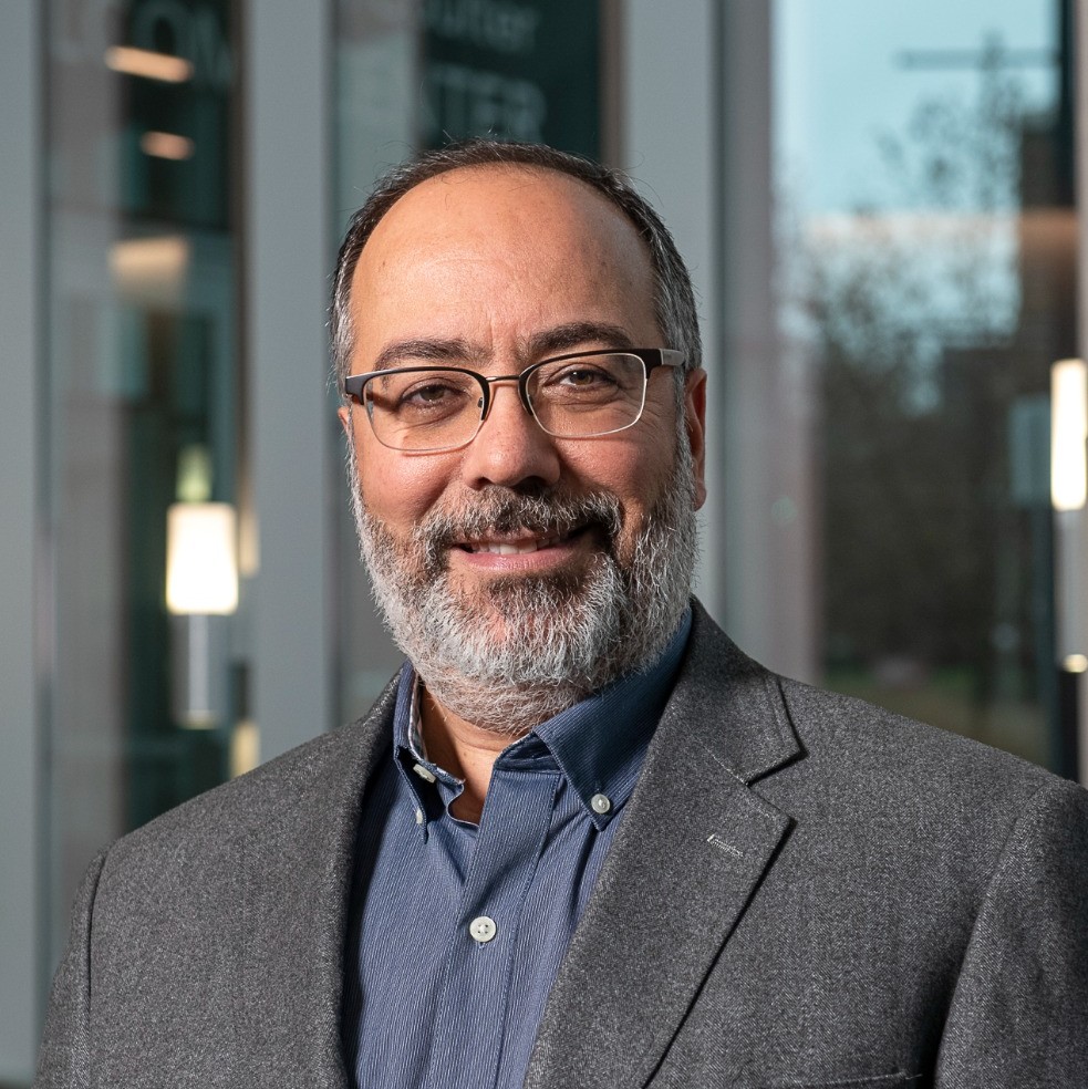 Middle-aged man with glasses, gray beard, blue shirt, and gray blazer smiling in an indoor setting.