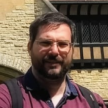 Man with glasses, beard, and backpack, standing in front of a brick and timber building.